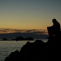 silhouette of man and woman sitting on rock near body of water during sunset